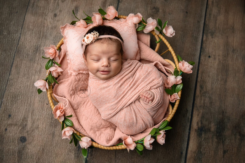 baby girl wrapped in peachy pink with toes showing, a floral headband, and a big smile on her face posed in a wicker basket with pink flowers all around it on a dark floor at her in-home newborn photo session in Houston, Texas.