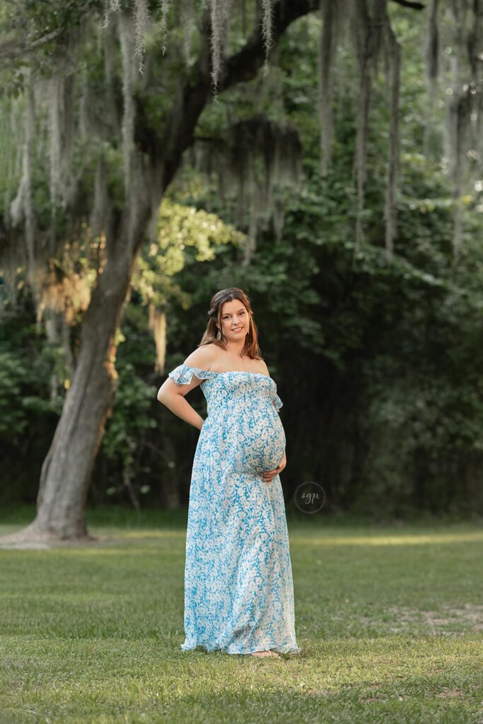 pregnant woman in long, blue floral dress under canopy of mossy trees at Houston outdoor maternity photo session