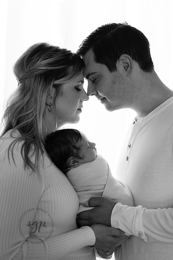 black and white portrait of new parents holding baby boy between them all dressed in cream all with eyes closed at Cypress in-home newborn photo session