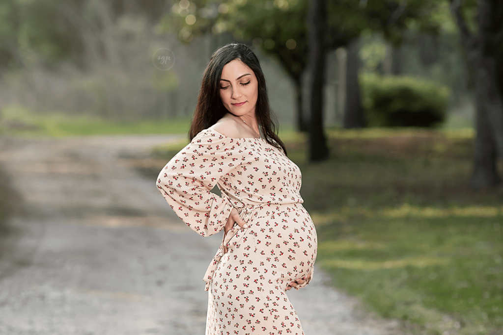 Mom to be in long cream and floral dress showing off baby bump at outdoor maternity photoshoot in Cypress, Texas.