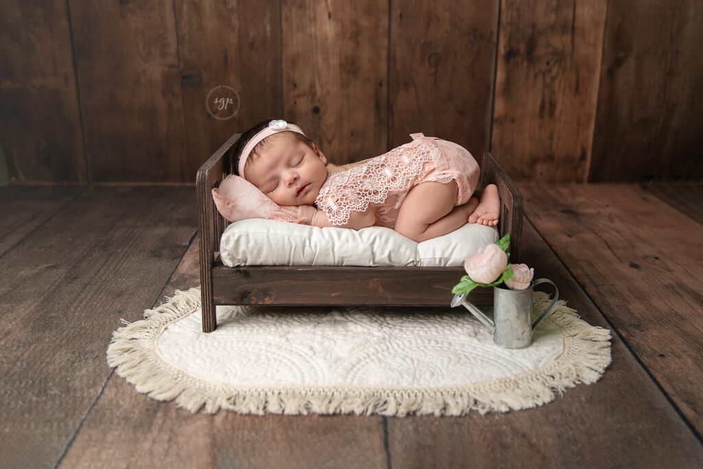 baby girl wearing pink lace onesie sleeping on tummy on a baby bed with some pink flowers and dark wood backdrop at in-home newborn photo session in Houston Texas.