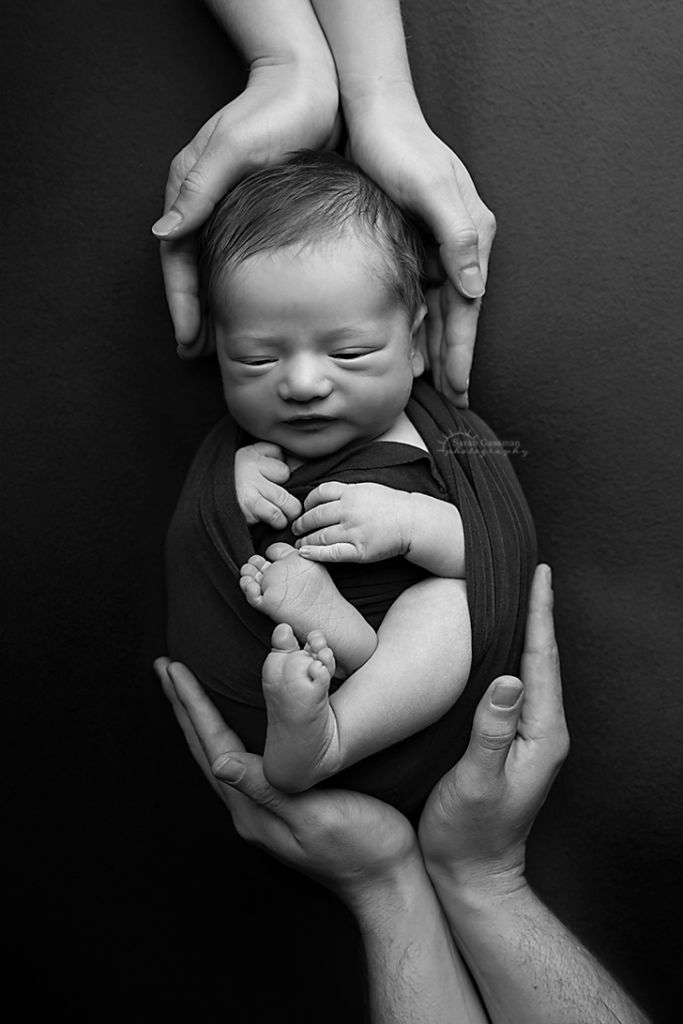 baby in a dark wrap being snuggled by his parents hands around him during his Cypress Texas in-home newborn photoshoot with Houston newborn photographer