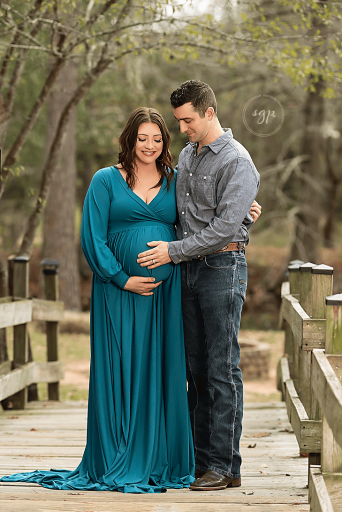 Couple embraces at outdoor maternity photo session with mom to be in long teal dress with dad looking lovingly at baby bump in Cypress, Texas.