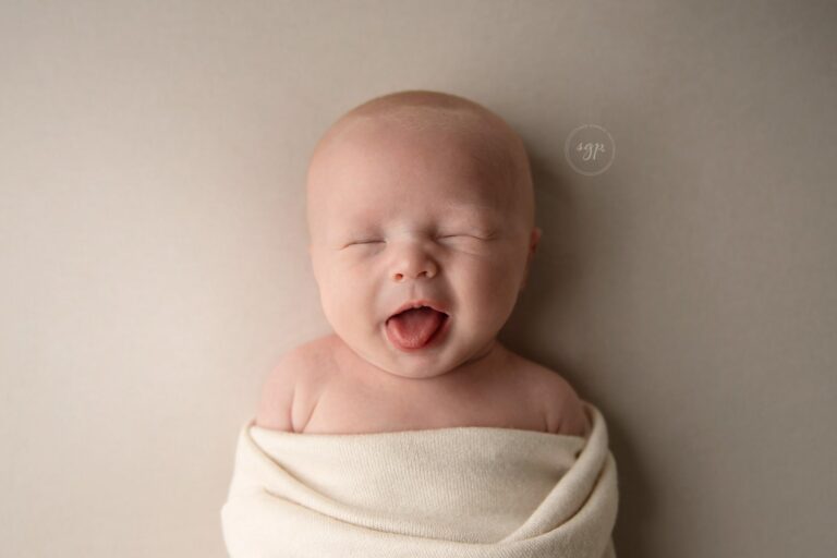 newborn baby making silly face with tongue sticking out with a cream wrap and backdrop at Houston newborn photoshoot in his own home