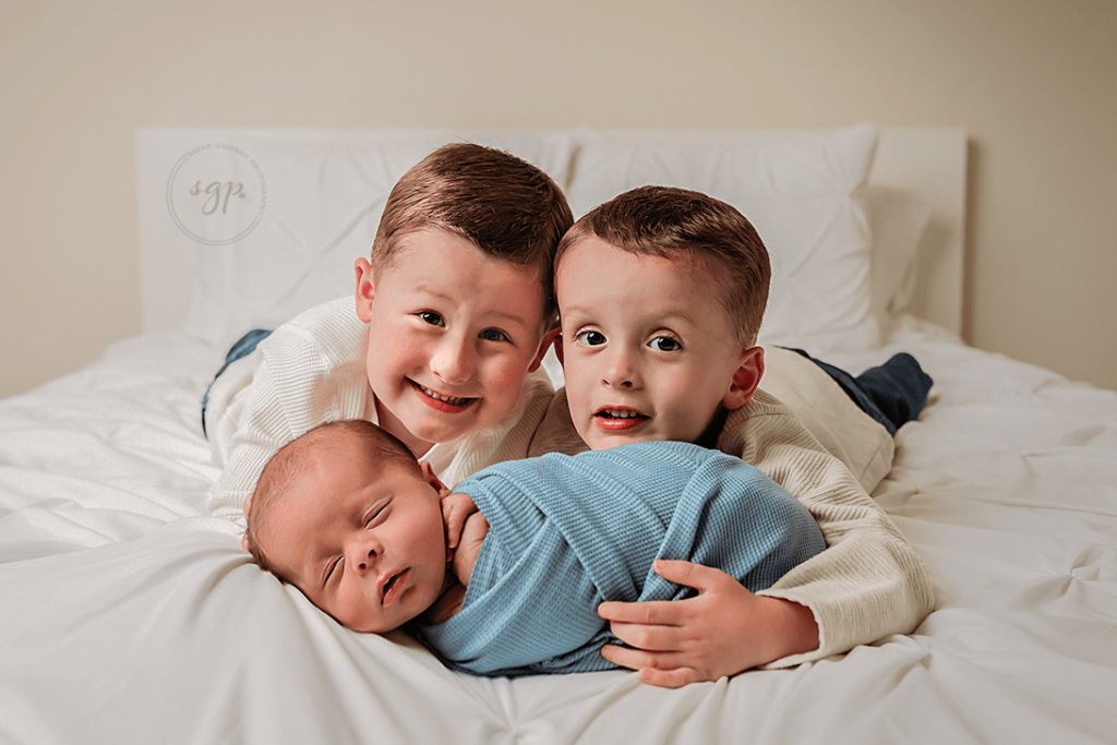 bog brothers happily hold their new baby brother on a bed in their own home for their Houston in-home newborn photo session