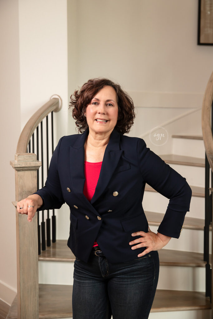 realtor in a navy jacket and pink shirt posing for headshots on stairs in a home in Cypress, Texas