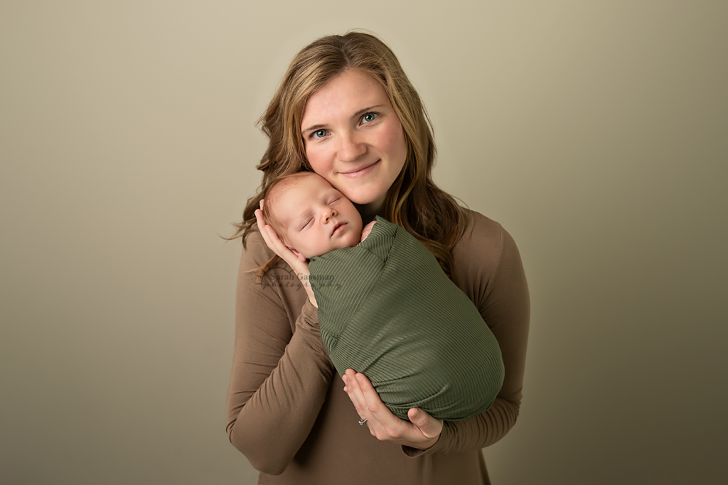 Mom dressed in brown dress holds sleeping baby boy wrapped in olive green at his in-home newborn photoshoot in Houston, Texas