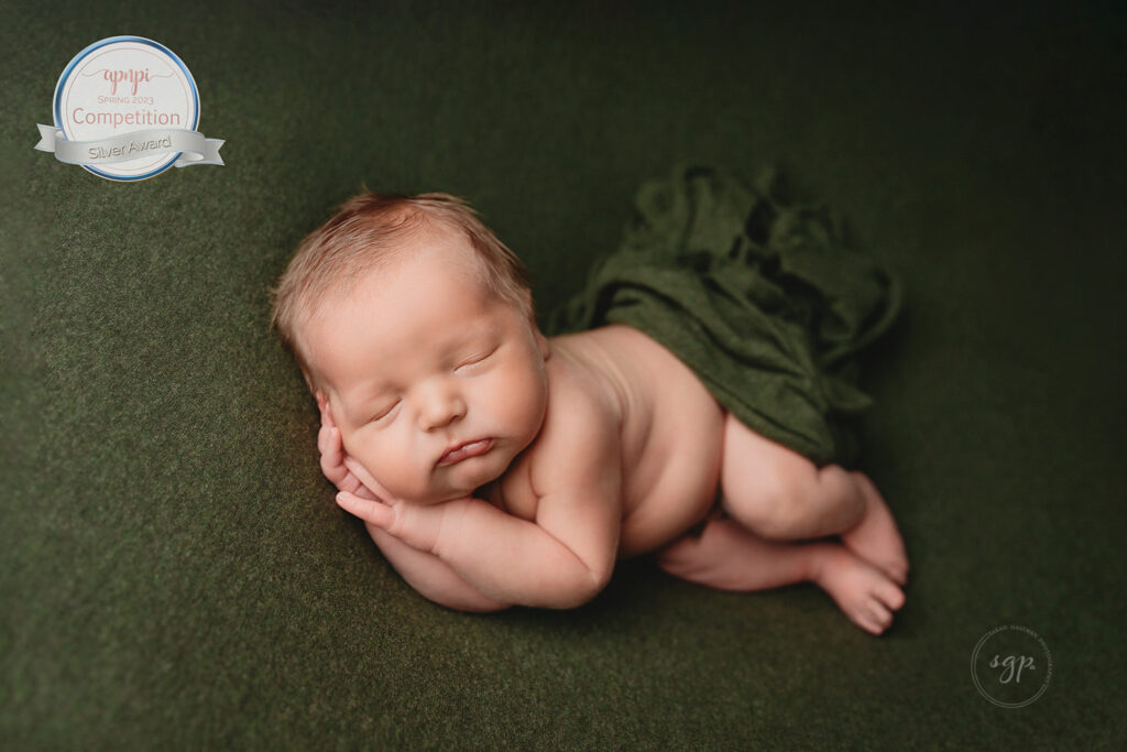 international award-winning photo of a baby boy sleeping on his side on a deep green backdrop with a matching green wrap covering his back half taken at his in-home newborn photo session by Houston newborn photographer