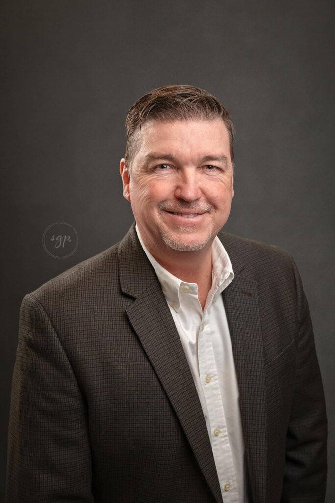 corporate headshot of man in white button up and brown jacket