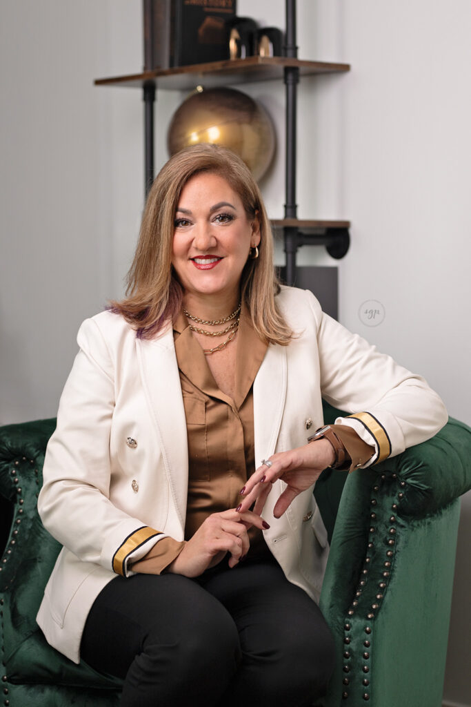 Woman wearing gold top with white jacket and black pants on green chair with gold accents in modern headshot