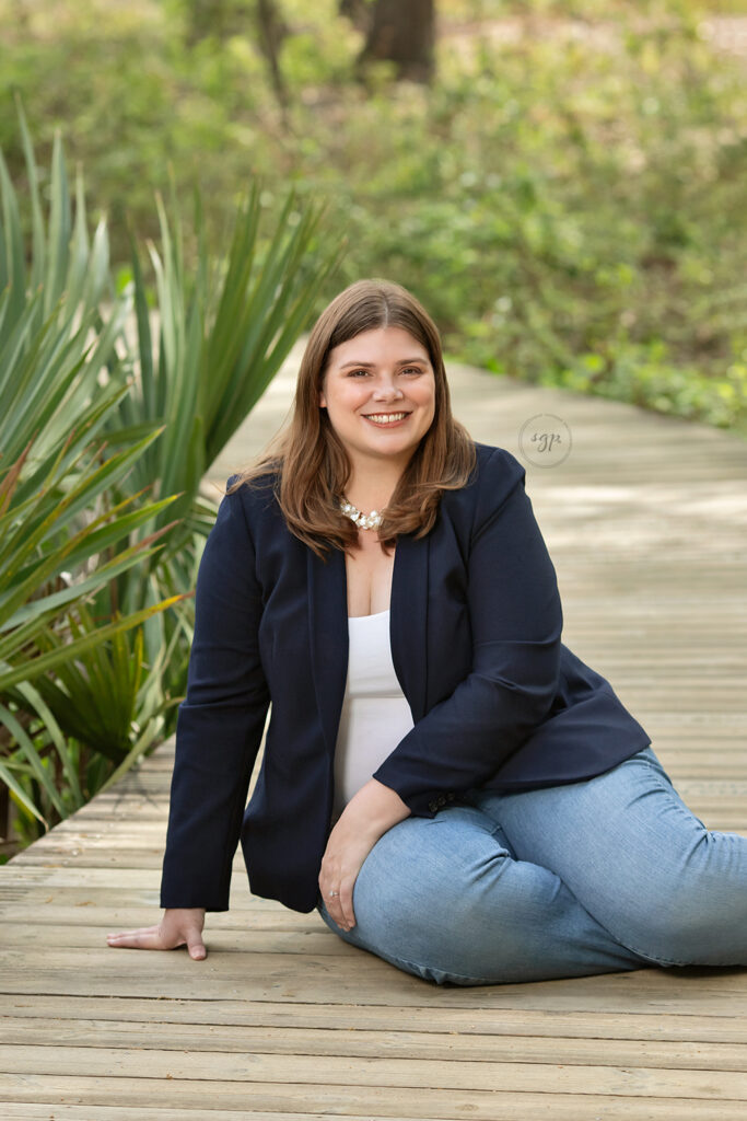 outdoor modern headshot of woman wearing jeans and blazer sitting on wooden path surrounded by greenery