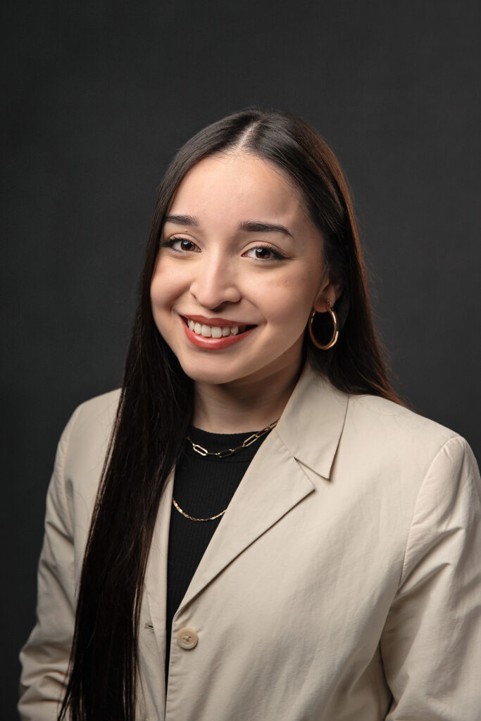 simple headshot of woman in greige blazer and black shirt with long, dark hair with a gray backdrop