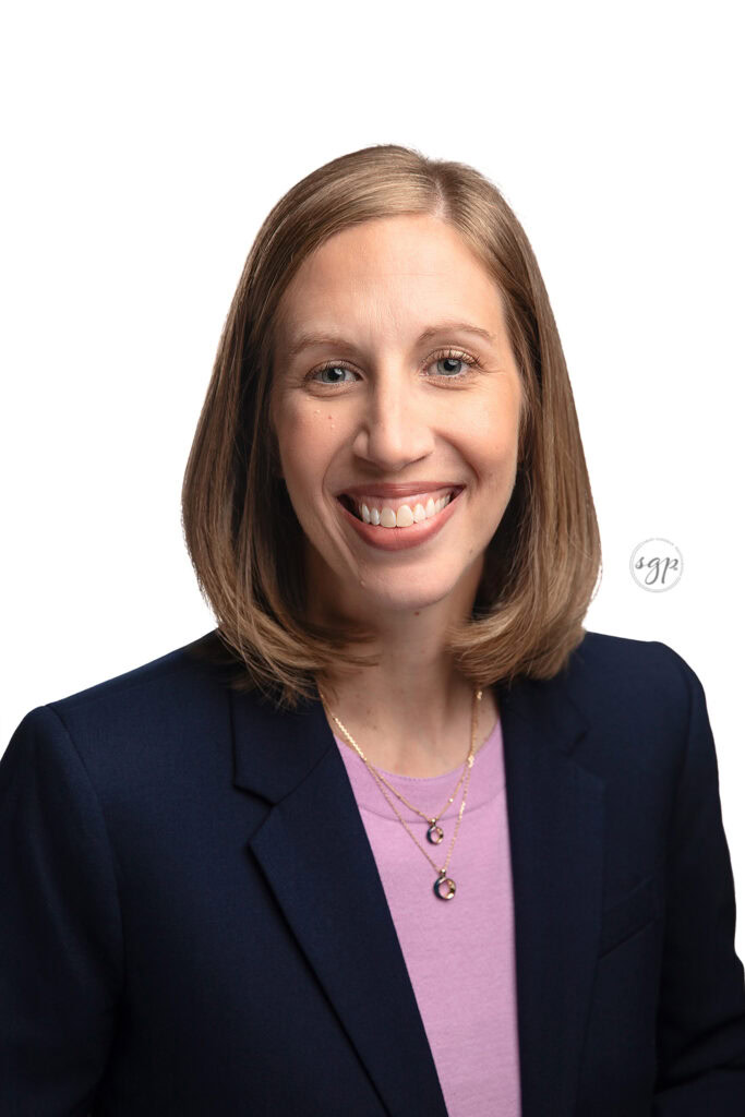 woman in lavender top with navy blazer smiles in corporate headshot with plain white backdrop